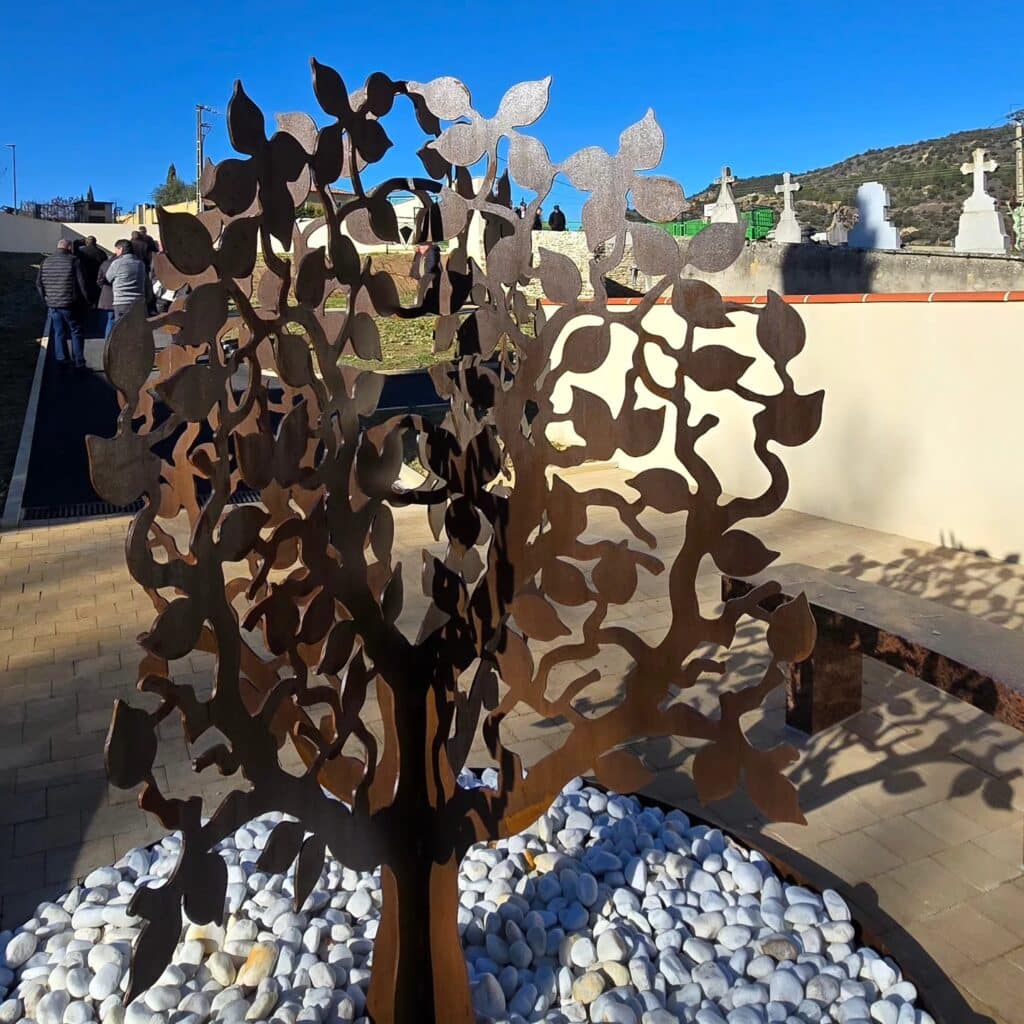 Sculpture d'arbre en métal découpé sur un lit de galets blancs dans un cimetière, mémorial symbolique.